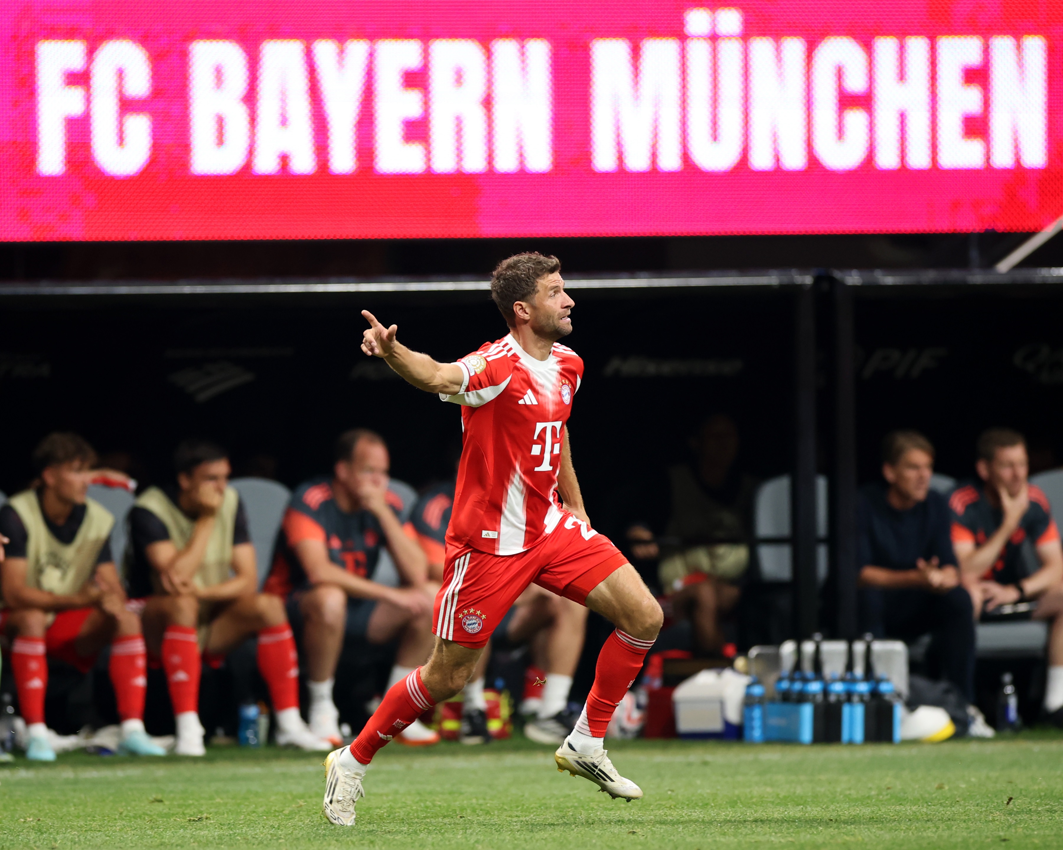 Thomas Muller runs on the pitch with Bayern teammates sitting on the bench in the background.
